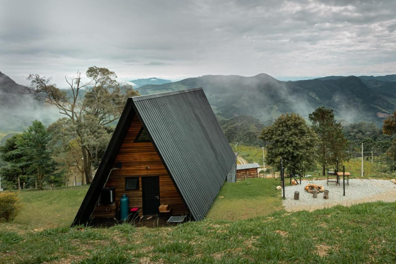 Una cabaña moderna de madera triangular con techo metálico, rodeada de césped y árboles, con montañas al fondo.