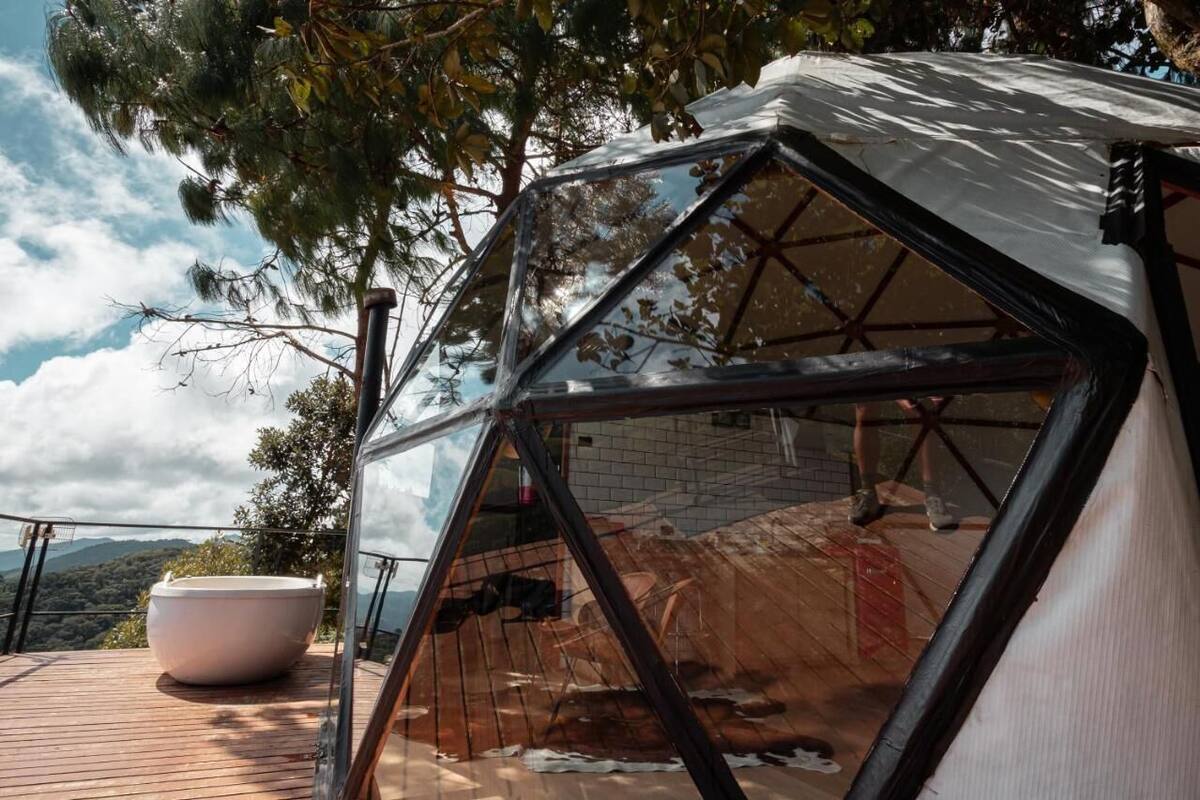 A geodesic dome structure with a transparent roof, a large white bathtub, and a wooden deck overlooking a mountainous landscape.