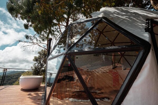 Una estructura en forma de cúpula geométrica con techo transparente, una bañera blanca grande y una terraza de madera con vistas a un paisaje montañoso.