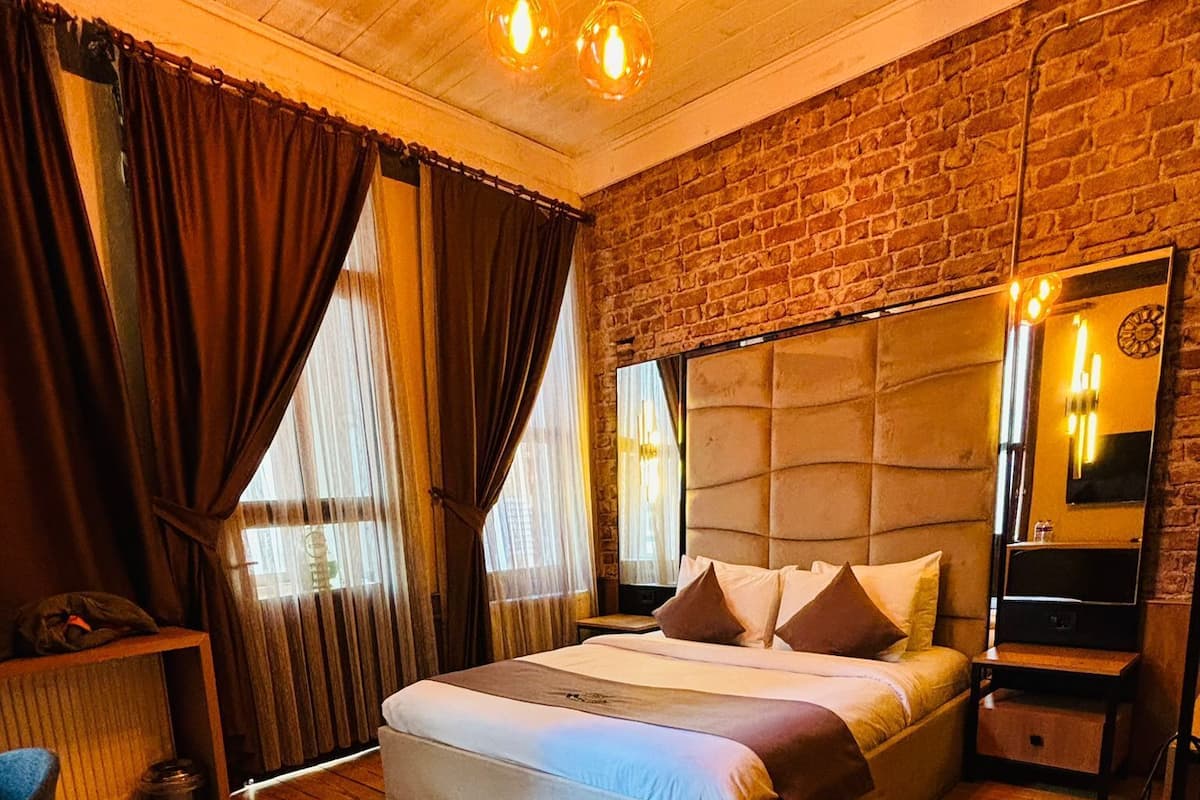 A bedroom with a wooden floor, a bed with pillows, a nightstand, a mirror, and a brick wall.