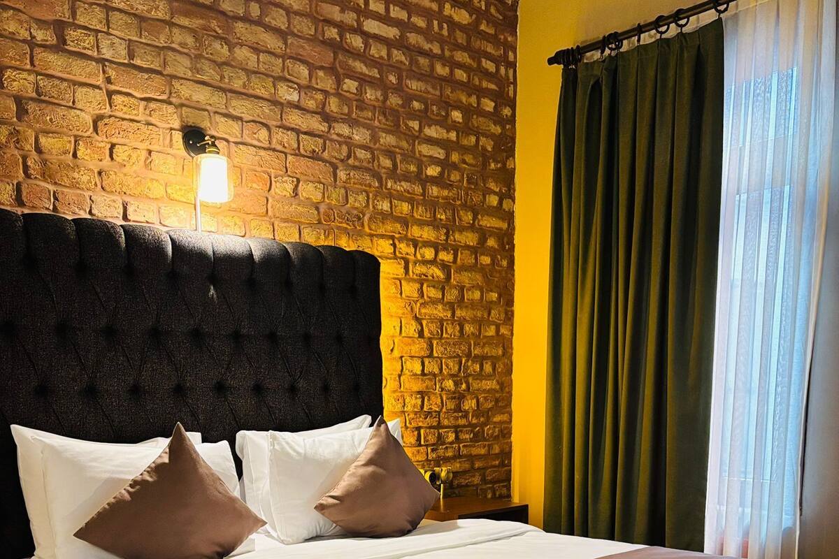 A hotel room with a brick wall, a large bed with multiple pillows, a bedside table, and a window with curtains.