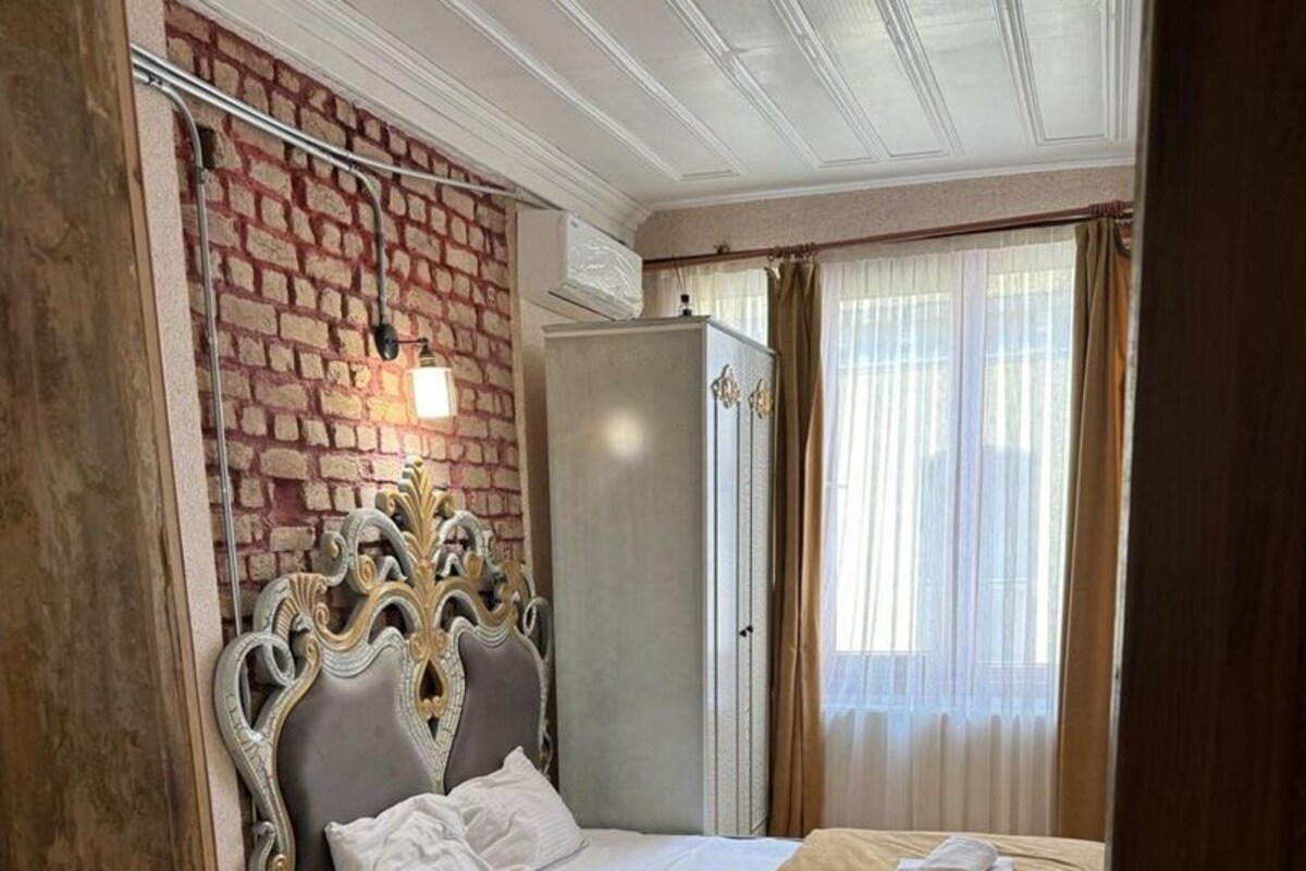 Una habitación con cama, armario, una mesita, espejo y pared de ladrillo.