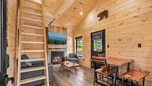 Un intérieur de chalet chaleureux avec une cheminée, un coin repas et un lit en mezzanine.