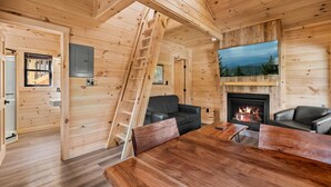 A wooden cabin interior with a fireplace, a dining table, a sofa, and a ladder.