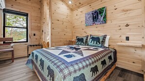 Une chambre dans une cabane en bois, avec un lit, une fenêtre, une chaise et une couverture à motifs d’ours.