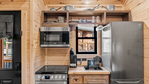 A compact kitchen with a microwave, stove, refrigerator, and wooden shelves with cookware.