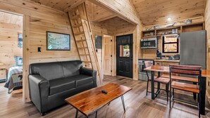 A wooden cabin interior with a sofa, coffee table, dining table, chairs, kitchen area, and a ladder.