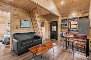 Interior de una cabaña de madera con sofá, mesa de centro, mesa de comedor, sillas, zona de cocina y una escalera.