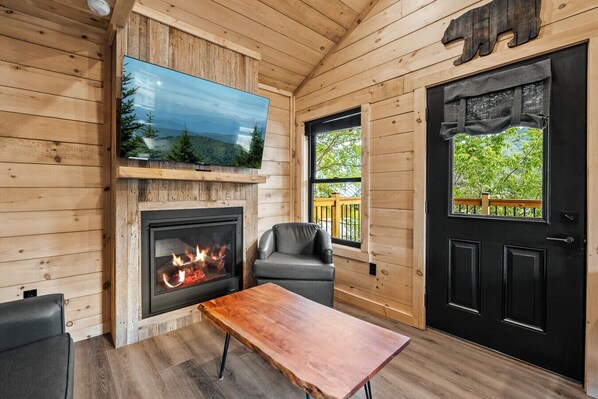 A cozy cabin interior with a fireplace, a TV mounted above it, a dining table, and a door with a view of the outdoors.