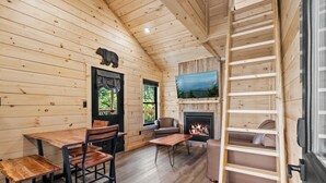 Un intérieur de chalet chaleureux comprenant un coin repas, une cheminée, un téléviseur et un lit en mezzanine.
