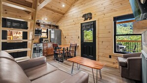 Un intérieur de chalet chaleureux comprenant un coin repas, une cuisine et une grande fenêtre donnant sur le paysage extérieur.