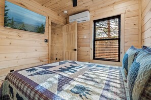 A wooden cabin interior with a bed, a window with blinds, a painting on the wall, and an air conditioning unit. - Little Forge TreeLodge, Downtown Pigeon Forge (Pigeon Forge)