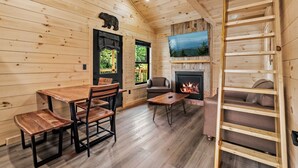 L’intérieur d’un chalet en bois comprend une table à manger, des chaises, un canapé, une cheminée, un téléviseur fixé au-dessus de la cheminée et une échelle en bois.
