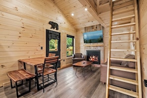 Interior de una cabaña de madera con una mesa de comedor, sillas, un sofá, una chimenea, un televisor montado sobre la chimenea y una escalera de madera.