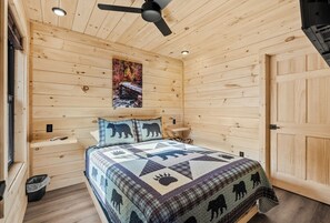 Une chambre dans une cabane en bois, avec un lit, un ventilateur de plafond, un tableau et une porte en bois.