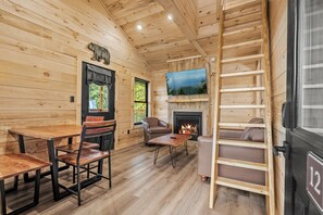 Interior de una cabaña de madera con chimenea, televisor montado encima y una cama en altillo con escalera.