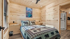 A wooden cabin bedroom with a bed, pillows, a ceiling fan, a painting, and a shower.