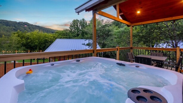A hot tub with a wooden deck and a table, surrounded by trees and a view of hills.