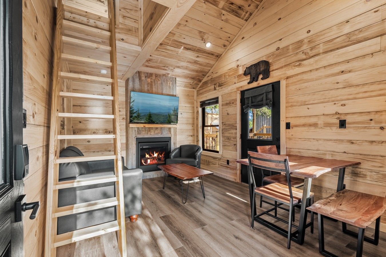 A cozy cabin interior with a fireplace, wooden walls, a TV mounted above it, and a dining area with chairs and a table.