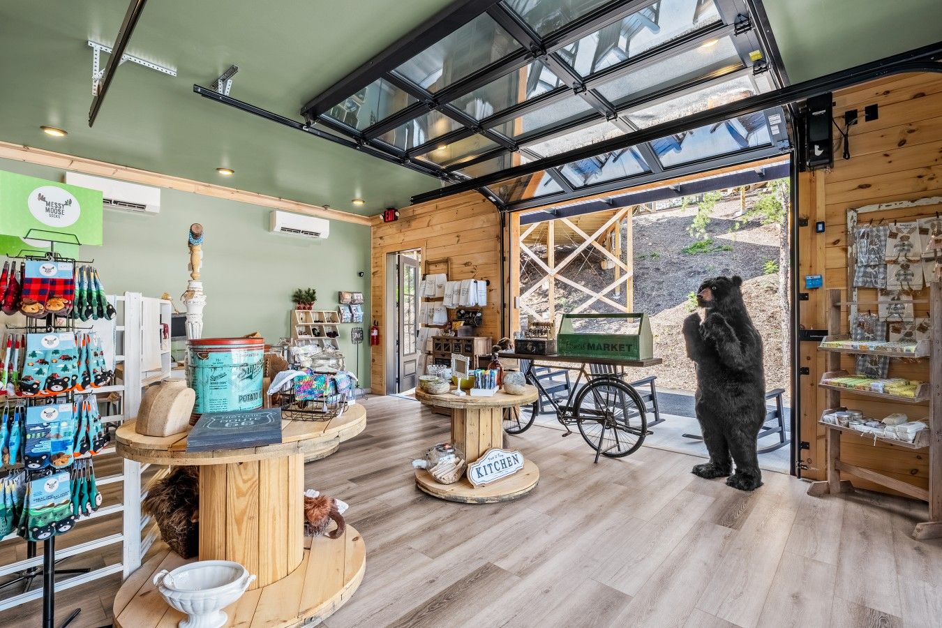 A rustic interior with wooden floors, a glass ceiling, and a variety of items on display including kitchenware and a bear statue.