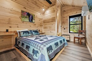 A wooden cabin bedroom with a bed, a chair, a window, and a bear-themed quilt.