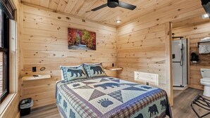A wooden cabin bedroom with a bed, a ceiling fan, a painting, a sink, and a shower.