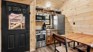 Kompakti keittiö, jossa on mustat ovet, puuseinät, ruostumattomasta teräksestä valmistettu jääkaappi, uuni ja ruokapöytä tuoleineen.