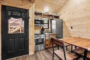 A compact kitchen with a black door, wooden walls, a stainless steel refrigerator, an oven, and a dining table with chairs.