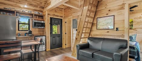A cozy cabin interior with a kitchen, dining area, and living room, featuring wooden walls and a black leather sofa.