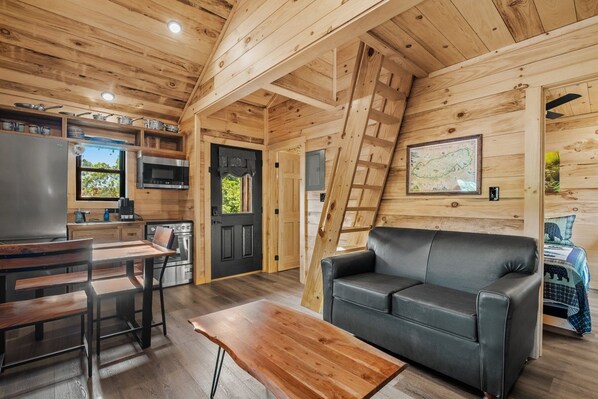 A cozy cabin interior with a kitchen, dining area, and living room, featuring wooden walls and a black leather sofa.