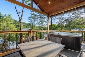 A wooden deck with a hot tub, a table, and chairs, overlooking a body of water and surrounded by trees. - TreeLodge, Downtown Pigeon Forge (Pigeon Forge)