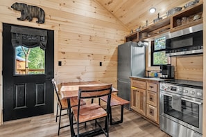 A wooden cabin interior with a dining table, chairs, a refrigerator, a stove, and a window with a view of trees. - TreeLodge, Downtown Pigeon Forge (Pigeon Forge)
