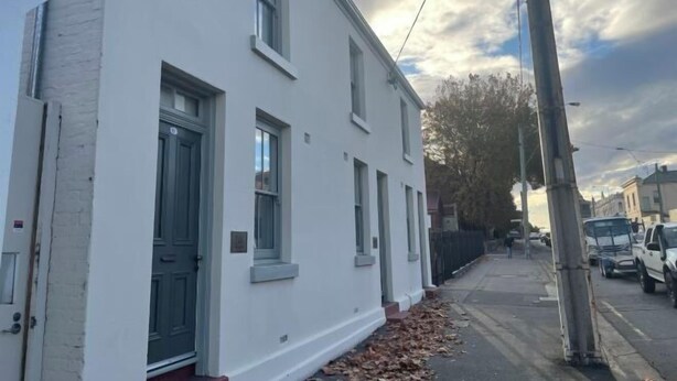 A white two-story building with a dark green door and windows, a sidewalk with fallen leaves, and parked cars along the street. - Urban Nook Serviced Apartments (Launceston)