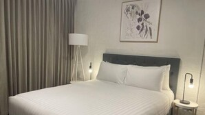 A neatly made bed with white linens, a dark headboard, a bedside lamp, and a framed artwork on the wall. - Urban Nook Serviced Apartments (Launceston)