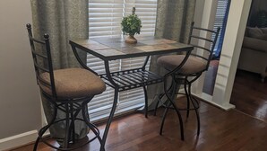 Un coin repas comprenant une table et deux chaises, une plante en pot et une fenêtre avec des stores.