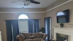 Un salon doté d’une cheminée, d’un ventilateur de plafond et d’une grande fenêtre avec des rideaux.