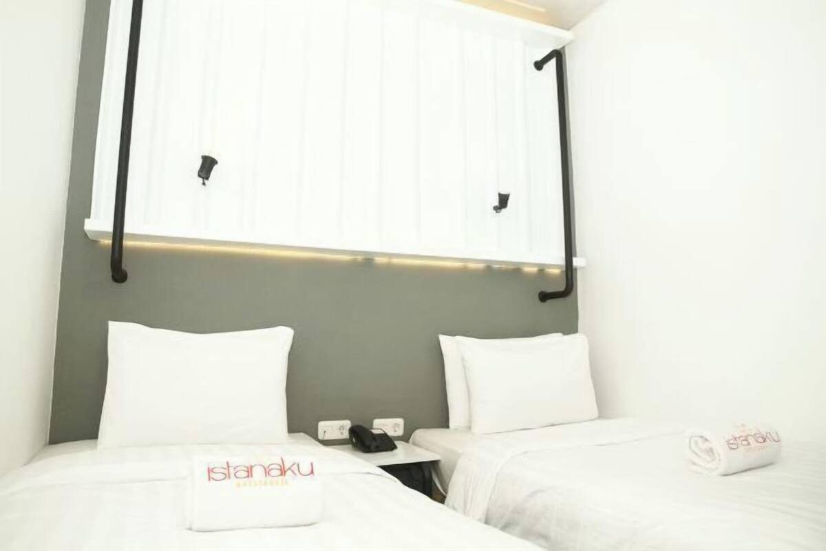 Habitación de hotel con dos camas, cabecera blanca, mesita de noche pequeña con teléfono y lámpara, y toalla con el nombre del hotel.