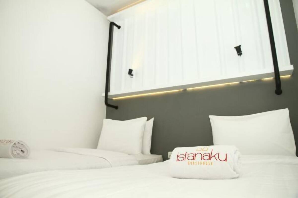 Habitación de hotel con cama, ropa de cama blanca, cabecera con iluminación integrada y almohada con el nombre del hotel.