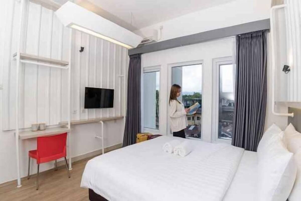 Habitación de hotel con cama, escritorio, silla, televisor y balcón con cortinas.