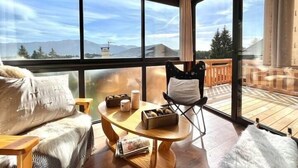 A cozy room with a wooden ceiling, large windows, a round wooden table, a chair, and a sofa with cushions.