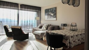 Una sala de estar moderna con un sofá, dos sillones, una mesita de centro y una mesa de comedor con mantel estampado. Hay un cuadro de ciervos en la pared, una lámpara y vistas al exterior a través de grandes ventanales.