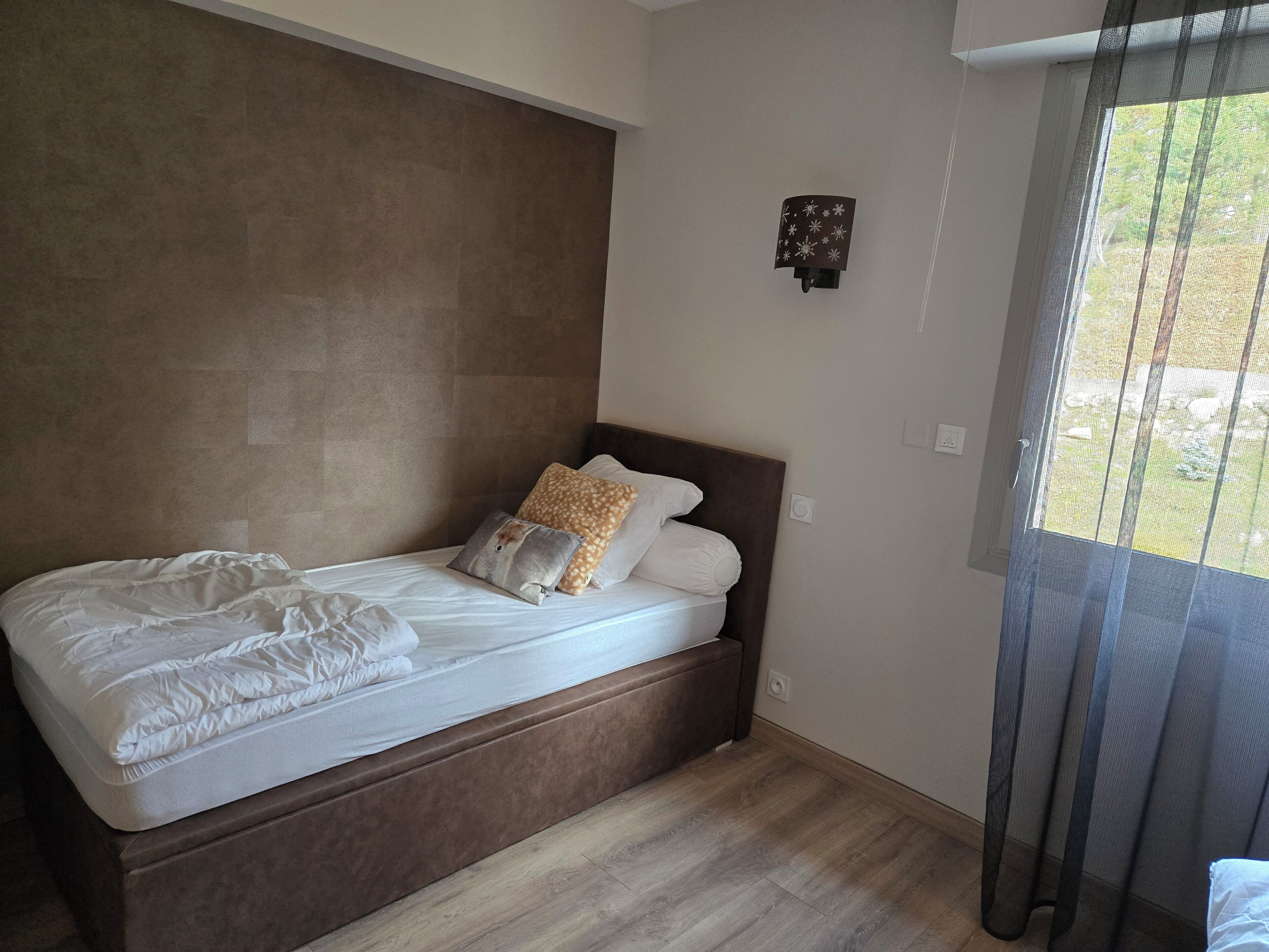 A bedroom with a bed, a headboard, a wall-mounted light fixture, and a window with curtains.