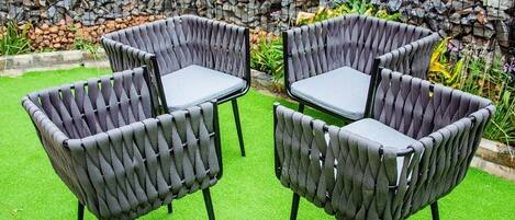 A set of four grey woven chairs with black legs, arranged around a small table on a green artificial grass surface, with a stone wall and plants in the background.