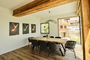 Un coin repas avec une table et des chaises en bois, des œuvres d’art représentant des cerfs encadrées au mur, et une vue sur un bâtiment à travers une grande fenêtre.
