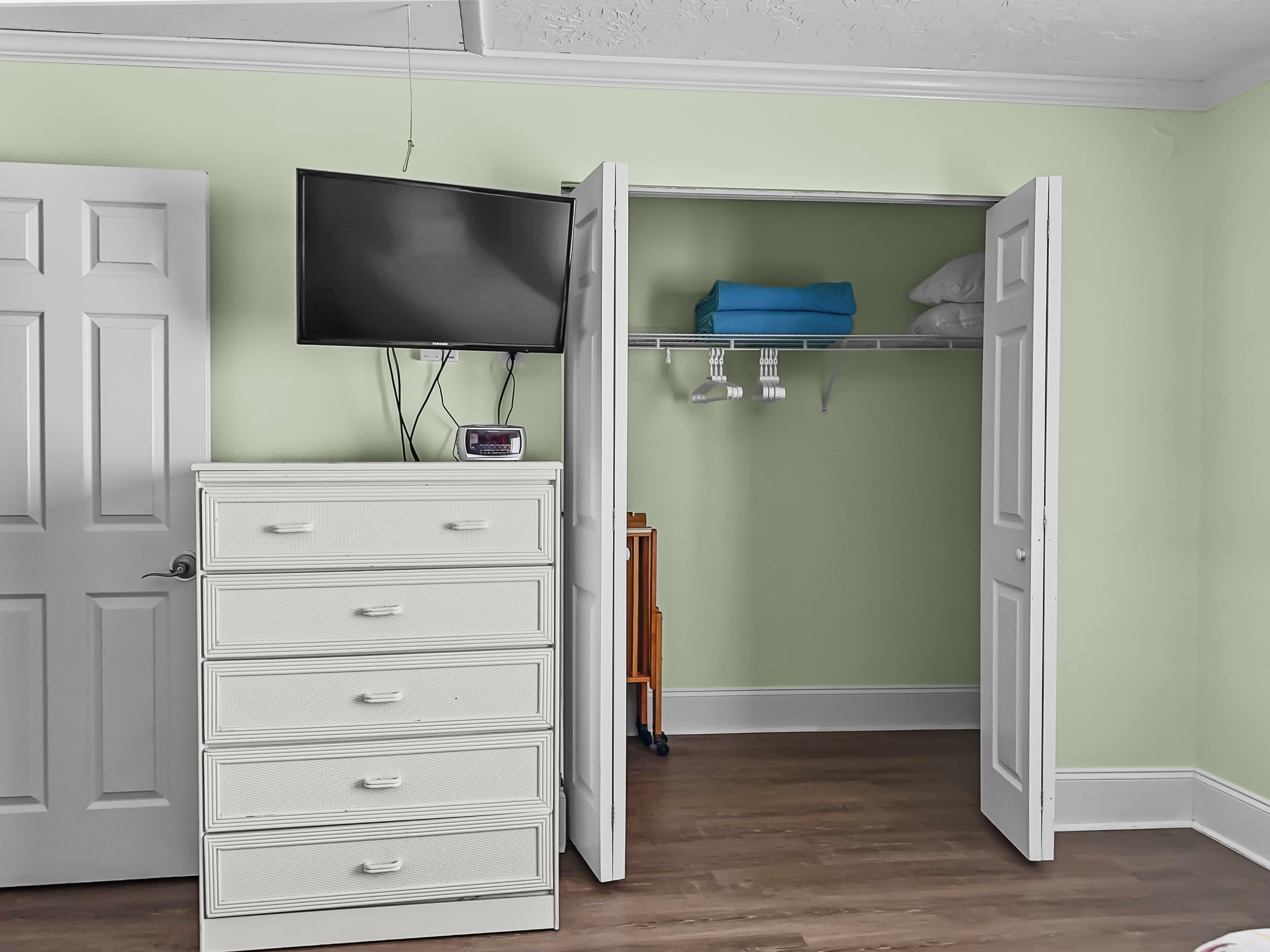 Ein Zimmer mit einem weißen Schminktisch, einem an der Wand befestigten Fernseher und einem Kleiderschrank mit blauem Handtuch und einem hölzernen Hocker.