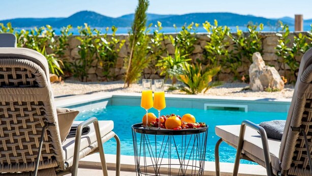 A poolside setting with lounge chairs, a table with fruit and drinks, and a view of the sea and mountains.