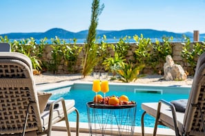 A poolside setting with lounge chairs, a table with fruit and drinks, and a view of the sea and mountains.