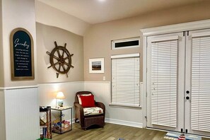 Una habitación con una silla de mimbre, una mesita con una lámpara, una decoración de timón de barco y una cita enmarcada en la pared.