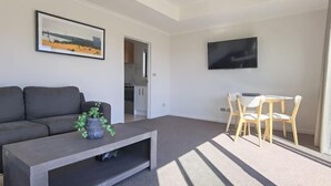 A modern living room with a grey sofa, a coffee table with a plant, a TV on the wall, and a dining area with wooden chairs. - RodmanStar Apartment (Lake Tekapo)