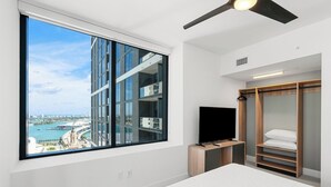 Una habitación de hotel moderna con un ventanal grande que ofrece vistas a la ciudad, una cama, un televisor sobre un mueble de madera y un vestidor.